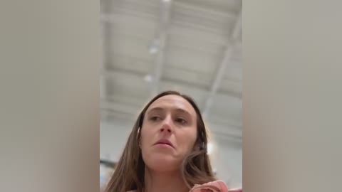 Video of a young woman with long brown hair, wearing a pink shirt, looking slightly annoyed, taken from a low angle inside a modern, white ceiling-tiled room.