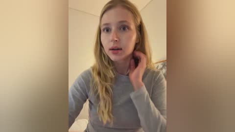 A video of a young, fair-skinned woman with long, wavy blonde hair, wearing a light grey long-sleeve top, standing in a dimly lit room with beige walls.