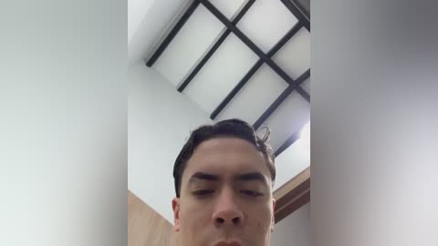 Video of a young Asian man with short, dark hair, looking down with a neutral expression, standing in a modern room with a grid-patterned skylight ceiling.