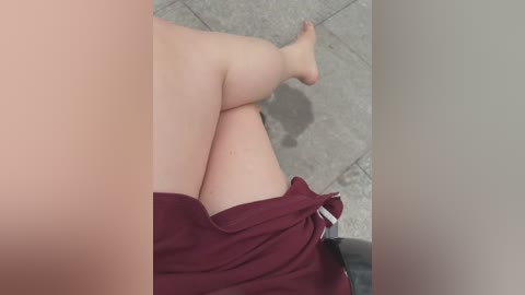 Video of a person's legs and thighs, wearing maroon shorts, lying on a grey tiled floor, barefoot. The image is close-up, with a blurred, out-of-focus background.