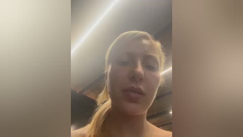 A video of a young woman with blonde hair tied back, standing in a dimly lit, possibly wooden, room. She has a neutral expression and is topless, partially obscured by the image's soft focus.