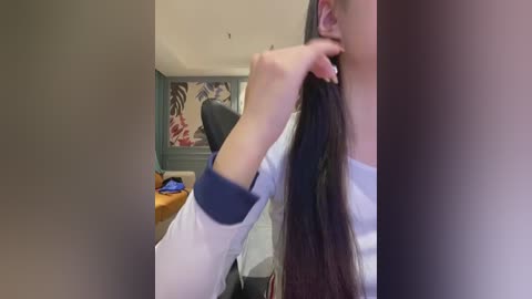 Video of a woman with long, dark hair, wearing a white blouse, using her left hand to pull her hair back. Background includes a green wall with floral art, a chair, and a desk with a laptop.