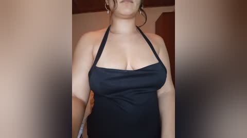 Video of a light-skinned woman with dark hair in a bun, wearing a black halter dress that accentuates her cleavage, standing in an indoor setting with neutral-colored walls.