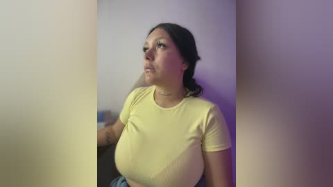 Video of a Latina woman with medium skin tone, wearing a tight yellow t-shirt that accentuates her large breasts and blue jeans. She stands indoors against a blurred, purple-toned background.