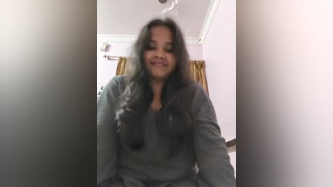 Video of a South Asian woman with long black hair, medium skin tone, wearing a gray shirt, seated in a room with light purple walls, white crown molding, and golden curtains.