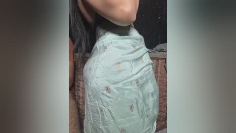 Video of a woman's bare back and shoulder, partially covered with a light blue sheet featuring subtle floral patterns. The background shows a dark, textured wall and a woven basket.
