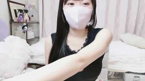 Media: Video of an East Asian woman with long black hair, wearing a white mask and black lace-trimmed top, standing in a cluttered, dimly lit room with white bed linens and shelves filled with personal items.
