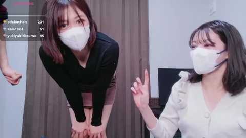 A video of two East Asian women in face masks, one bent over, the other smiling, in a dimly lit room with a wooden door.