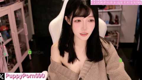 Video of a young Asian woman with long black hair, fair skin, and small breasts, wearing a beige off-shoulder sweater, sitting on a white gaming chair in a cluttered room.