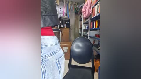 Video of a cluttered room with a person wearing a red shirt and blue jeans, sitting on a black chair, surrounded by stacks of boxes, a pink bag, and shelves filled with books and items.