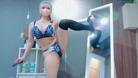 Video of a blonde woman in blue lingerie and a headband, balancing on one leg, holding a black phone, in a modern, bright room with shelves and mirrors.