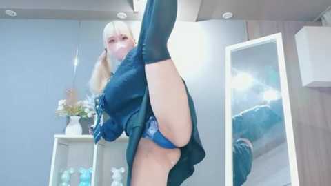 A video of a blonde woman in a green outfit, lifting her leg high, in a modern room with white furniture and a mirror.
