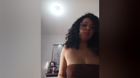 Video of a woman with curly black hair and glasses, wearing a strapless brown dress, standing in a dimly lit room with a window and a doll on a shelf.