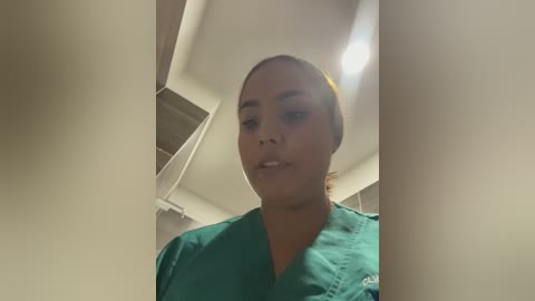 Video of a light-skinned woman with medium-length brown hair tied back, wearing green scrubs, standing indoors with ceiling lights overhead.