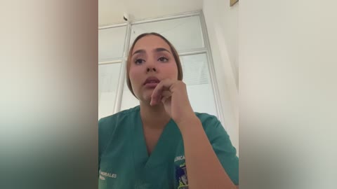 A video of a young woman with light skin and brown hair, wearing green scrubs, contemplating with her hand to her chin in a sterile medical setting with white walls and a window.