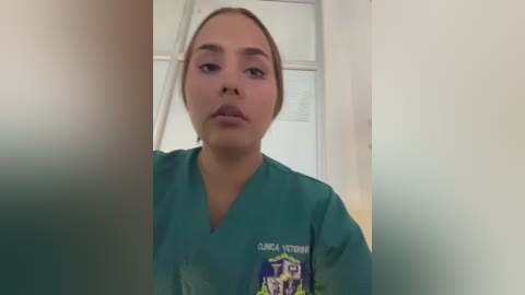 Video of a young, light-skinned Latina woman with straight brown hair, wearing teal scrubs with a badge. She gazes directly at the camera in a hospital setting.