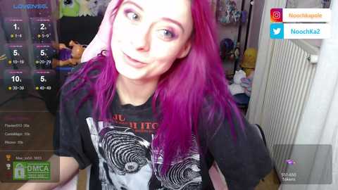 Media: Video of a young woman with vibrant pink hair and fair skin, wearing a black graphic t-shirt, smiling in a cozy bedroom with a digital overlay showing social media icons.