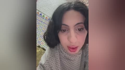 A video of a woman with fair skin and dark hair, wearing a polka-dot dress, standing in a room with a patterned rug and tiled floor.