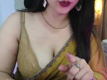 Video of a fair-skinned woman with long black hair, wearing a mustard yellow saree with a plunging neckline, large silver earrings, and a black necklace. Her lips are painted deep red.