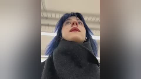 Video of a person with pale skin, blue hair, and dark clothing, seen from a low angle, looking upwards with a serious expression, in an indoor setting with white ceiling.
