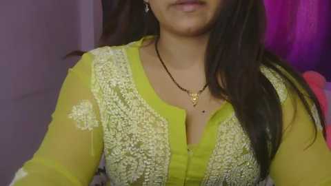 Media: Video of a South Asian woman with medium skin tone, wearing a bright yellow kurta with intricate white embroidery and a black necklace. She has long, dark hair and is smiling. Background shows purple curtains.
