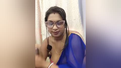 Video of a South Asian woman with medium skin tone, wearing a blue saree with gold trim, glasses, and a bindi. She holds a camera in her right hand, indoors, with a white textured wall in the background.