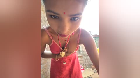 Media: Video of a young South Asian woman with short black hair, wearing a red sleeveless dress and red bindi, looking up from a low angle. She stands in front of a brick wall, with sunlight streaming in from the right.