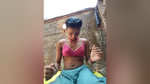 Video of a young South Asian woman with medium skin tone, braided hair, wearing a pink bra and green pants, sitting on a stone floor, appearing deep in thought.