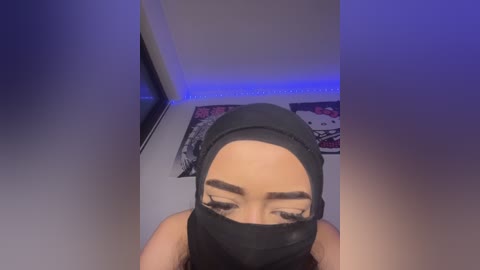 Video of a woman with medium skin tone and dark hair, wearing a black face mask and headscarf, in a dimly lit room with purple LED lighting, a Pikachu poster, and a TV in the background.