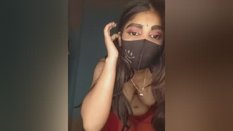 Media: Video of a woman with medium brown skin, long dark hair, and a black mask covering her nose and mouth, wearing a red top. She's indoors, adjusting her mask, with a blurred background.