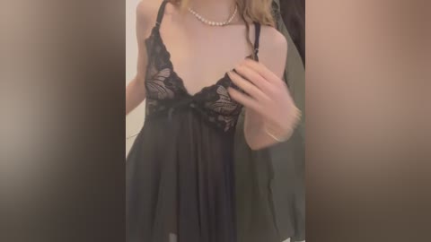 Video of a light-skinned woman wearing a sheer black lace babydoll nightgown with floral patterns. She has a pearl necklace and her long blonde hair is partially visible. Background is blurred, possibly indoor.