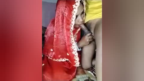 Video: Young Indian woman with dark skin and short hair in a red sari with white trim, performs oral sex on a man's erect penis. Background shows a yellow shirt.