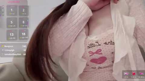 Media: A video shows a light-skinned woman with long brown hair, wearing a pink, polka-dotted top with a heart graphic and a white cardigan, in a modern, indoor setting.