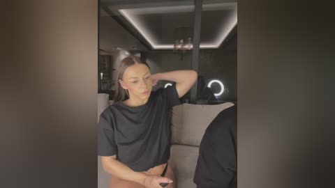A video of a light-skinned woman with brown hair, wearing a black crop top, sitting on a beige sofa in a modern, dimly lit room with a minimalist design.