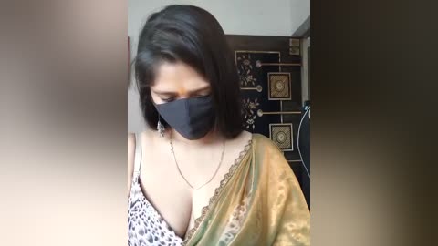 Media: Video of a woman in a leopard-print blouse, gold sari, black face mask, and gold earrings, standing indoors with a dark wall and ornate door behind her.