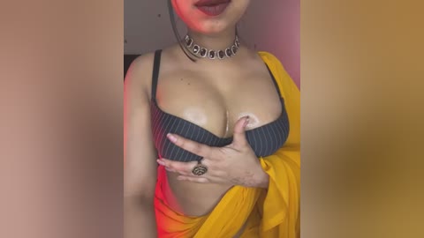 Video of a woman with medium skin tone, wearing a blue bra, yellow shawl, black choker, and a ring, partially covering her breasts. The background is a blurred gradient of orange and pink hues.
