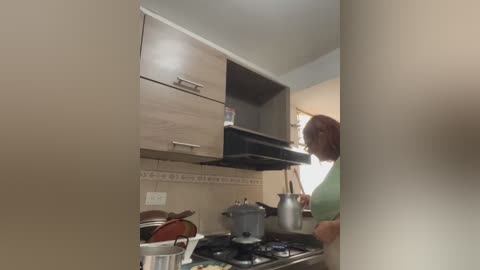 Video of a woman in a small kitchen with light wood cabinets, stainless steel appliances, and a gas stove; she's cooking with a pot, wearing a green shirt.