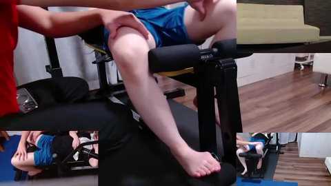 Media: Video of a young boy with light skin and fair hair, wearing blue shorts, being held and stretched on a black exercise machine in a modern gym with wooden floors and white walls.