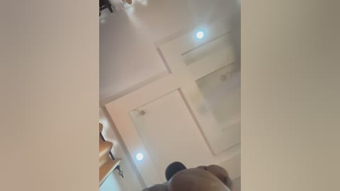 Video of a man with dark skin and a muscular build, naked, standing in a modern, white-walled room with recessed lighting and a wooden cabinet.