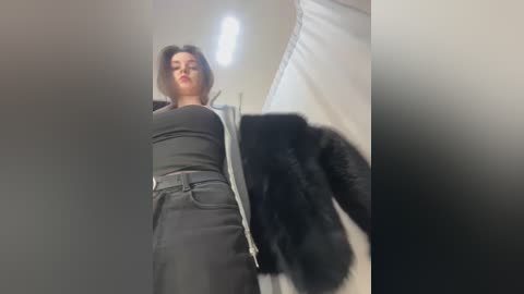 A video featuring a woman with short dark hair, dressed in a black top and high-waisted black skirt, standing in a dimly lit room with a black dog on the right.