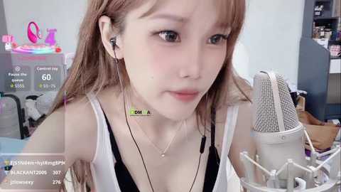A video of a young, pale-skinned, light-haired Asian woman with freckles and blue eyes, wearing a black bra, white camisole, and headphones, sitting in a cluttered room with a white microphone.