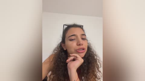 Video of a young woman with long, curly brown hair, wearing sunglasses, leaning on her elbow against a plain white wall, looking introspective.