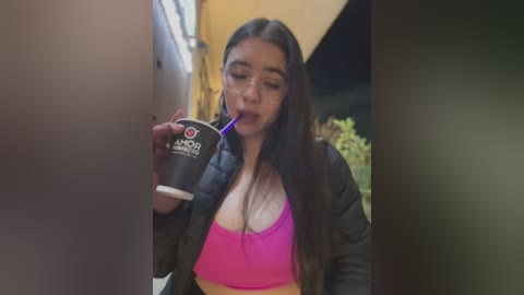 Media: Video of a young Latina woman with long dark hair, wearing a pink sports bra and black jacket, drinking from a black \"Torani\" coffee cup, standing outdoors near a wall and a window.