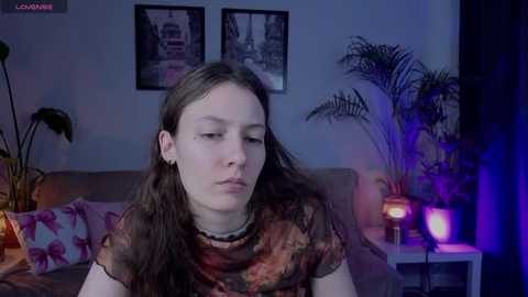 Video of a young woman with long brown hair, wearing a patterned blouse, seated in a dimly lit room with a potted plant, posters, and a lit lamp.
