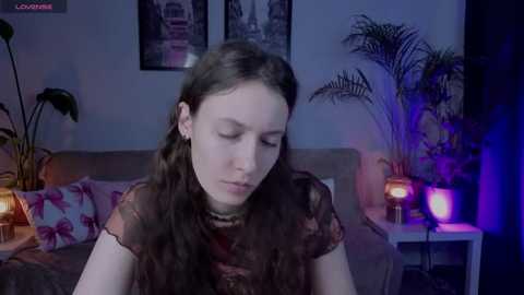 Video of a young woman with long brown hair, wearing a sheer black top, sitting on a bed in a dimly lit room with potted plants, framed pictures, and a lamp.