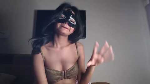 Video of a woman with long black hair, wearing a black blindfold, revealing her medium-sized breasts in a gold lace bra, with a neutral expression, in a dimly lit room.