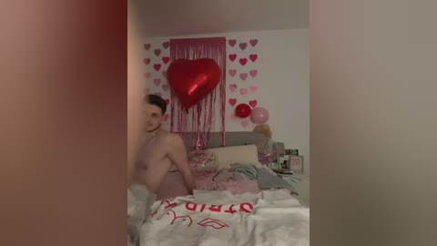 Media: Video of a young, shirtless man with fair skin and short dark hair, standing in a dimly lit, cluttered bedroom adorned with pink heart decorations and a large red heart-shaped balloon.
