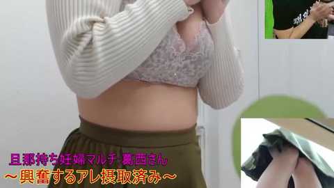 A video of a light-skinned woman in a beige knitted sweater lifting her shirt, revealing a lace bra, and olive green pants. The background includes a green object and a green and black shirt. Text in Japanese.
