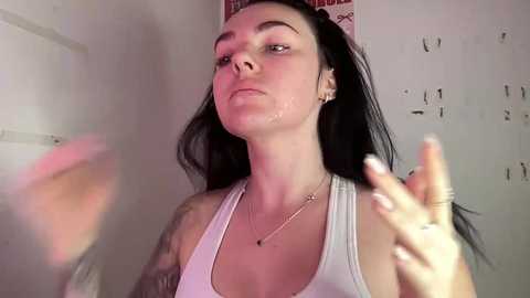 Video of a young woman with long black hair, wearing a white tank top, standing in a dimly lit room, wiping sweat from her face.