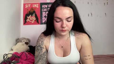 Video of a fair-skinned woman with long black hair, wearing a white tank top, with visible tattoos, standing in a bedroom with pink posters, a stuffed animal, and a bed.